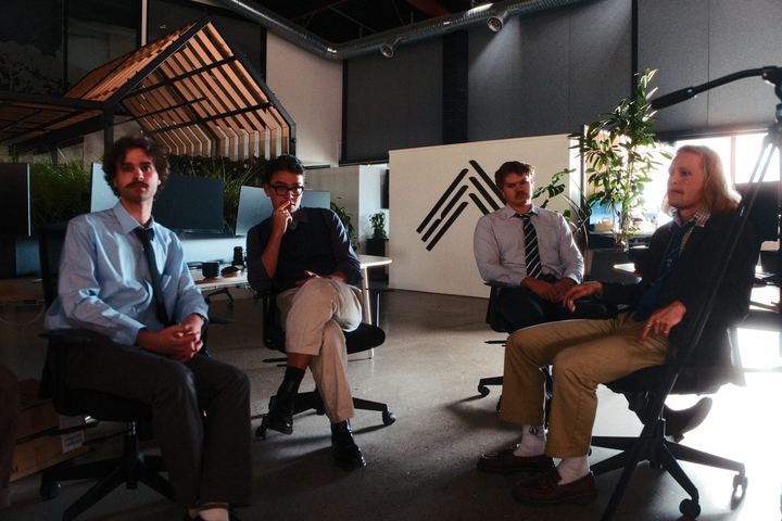 The band sitting in a circle of office chairs like a staff meeting