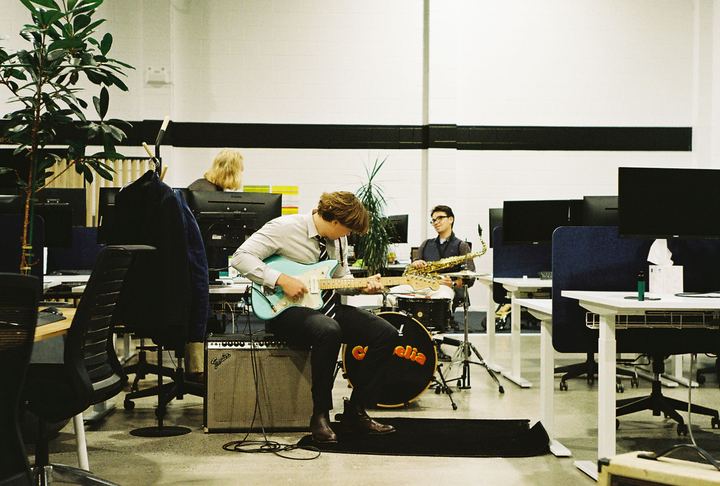 Guitarist on an amp playing a teal guitar between the office desks, sax player and drummer behind, shot on film